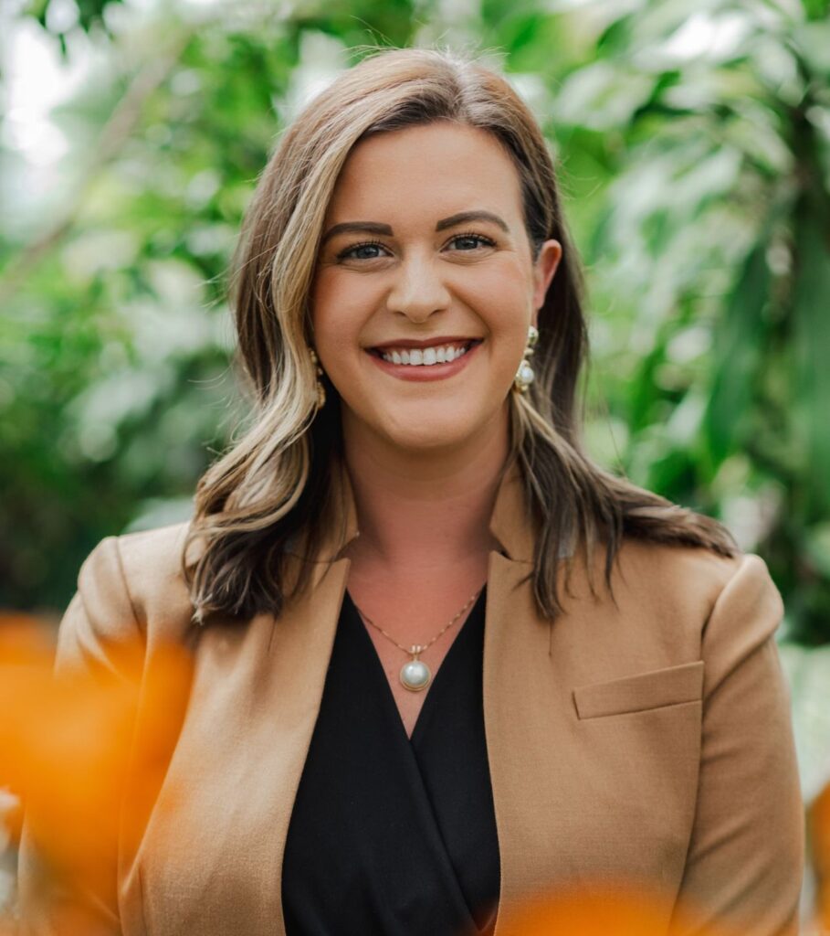 An individual in business casual dress standing in a natural setting and smiling at the camera.