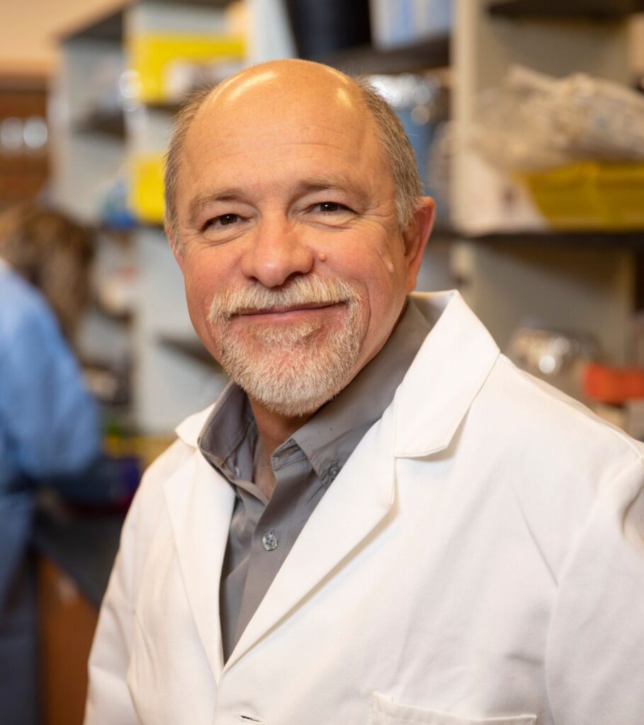 An individual wearing a labcoat in a research lab smiling at the camera.